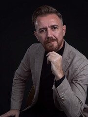 Portrait of adult businessman wearing trendy suit and sitting in modern studio on stylish chair against the black background