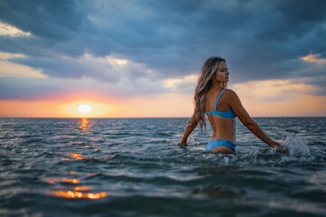 Fototapeta na wymiar A girl with blond hair in a blue swimsuit splashes to the sides while sitting in an estuary on a sunset background
