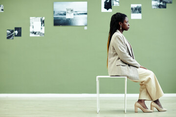 Side view of stylish young Black woman sitting on stool in modern art gallery looking at black and...