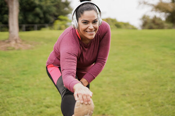 Young curvy woman doing sport stretching exercise at city park while listening music with wireless headphone - obrazy, fototapety, plakaty