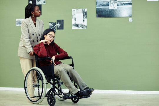 Young African American woman and her Asian male friend with disability in wheelchair visiting modern photography exhibition together