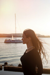 Silhouette of a strong confident woman on the seashore with a view of beautiful yachts. Portrait of a thin girl in a black dress at sunrise or sunset
