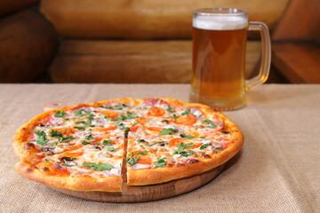 Traditional pizza with salami and champignons in front of a glass mug of light beer on the background of a log wall.