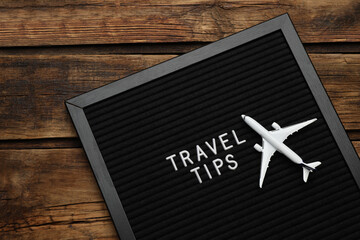 Black letter board with phrase Travel Tips and toy plane on wooden table, top view