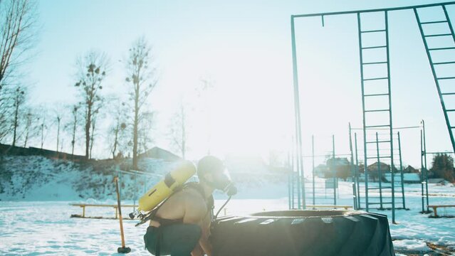 A muscular man with a naked torso with ballon compressed air drager on his back is doing crossfit and turn over tire. Slow mption