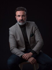 Portrait of adult businessman wearing trendy suit and sitting in modern studio on stylish chair against the black background