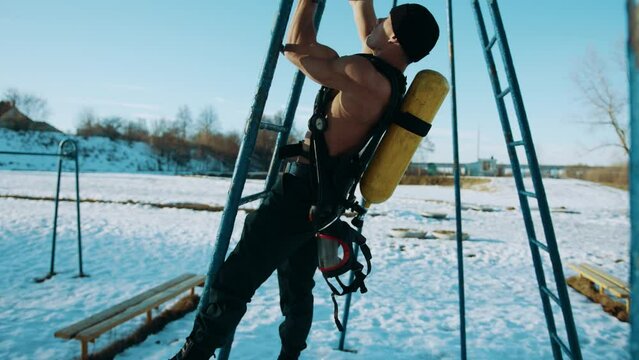 A muscular man with a drager on his back climbs on his hands up an inclined ladder on a sports field in a winter park