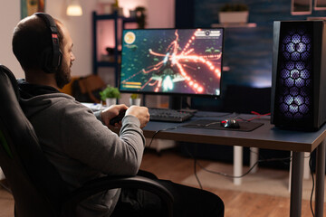 Naklejka premium Gamer using controller to play online video games on computer. Man playing game with joystick and headphones in front of monitor. Player having gaming equipment, doing fun activity.
