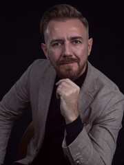 Portrait of adult businessman wearing trendy suit and sitting in modern studio on stylish chair against the black background