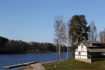 Fototapeta na wymiar Wooden house on shore of calm lake. Lake with pier in sunny spring day.