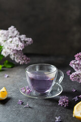 Romantic spring background with cup of purple tea and lilac flowers on dark background. Spring tea drinking. Greeting card, invitation design. Cafe menu, poster. Lilac tea recipe. Copy space
