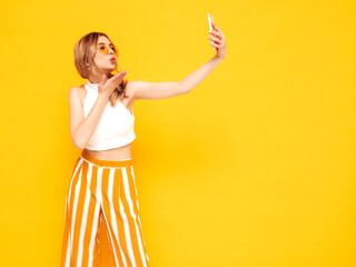 Portrait of young beautiful smiling blond female in trendy summer clothes. carefree woman posing near yellow wall in studio. Positive model having fun indoors. Cheerful and happy.Taking photo selfie