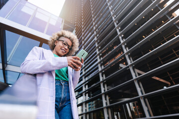 Trendy African American professional female using smartphone outside executive workplace. Creative businesswoman texting phone app