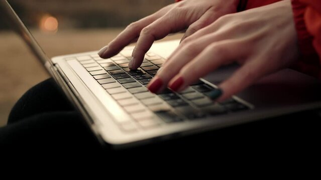Closeup shot of hands of a girl with painted nails typing on a laptop keyboard by the lake at sunset.