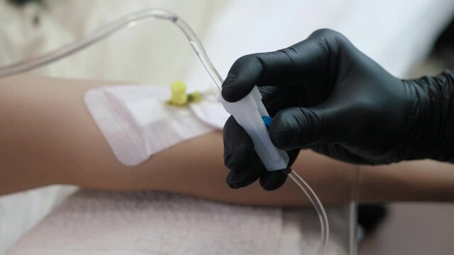 Doctor's hand in a black glove adjusts the medical drip valve. System for intravenous injections against the background of a hand with a catheter