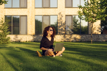 Cheerful young woman in sunglasses sitting on green grass with legs crossed during summer day while using laptop smiling.