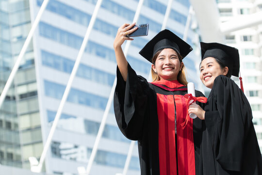 Two happy Asian young beautiful graduate female students with University degree standing and holding diploma taking selfie picture with mobile phone after graduation. Blur background of building