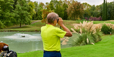 The golfer accurately measures the distance to the flag in a golf course with the laser rangefinder. In the background the forest.