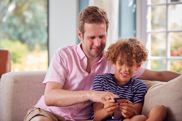 Father And Son Sitting On Sofa At Home Playing Game On Mobile Phone Together