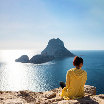 young woman in yellow zip jacket sitting on a cliff looking out on the Mediterranean Sea from Ibiza to famous Es Vedra