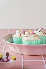 Three pastel colour easter egg cupcakes in a pink dish. Set against a pink tile worktop and plain background with copy space available. Mini chocolate eggs in a small bowl in foreground.