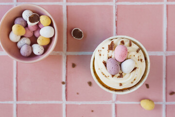 Top down view onto a Easter theme cupcake. Cake is decorated with flakes of chocolate and mini pastel colour eggs. Set against pink background. Spring light bright colour theme.