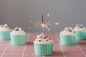 Easter egg cupcakes set against a pale pink tiled work surface and plain background. Cupcake decorated with mini chocolate eggs and cream frosting. Front cupcake has lit sparkler burning. - obrazy, fototapety, plakaty