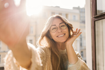 Pretty caucasian adult woman in glasses smiling while looking at camera while standing by open...