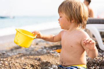 Cute child playing with yellow bucket and toy car on pebble beach. Happy toddler on sea coast. Summer family holiday vacation. Two years old little boy having fun. Summertime. Travel concept