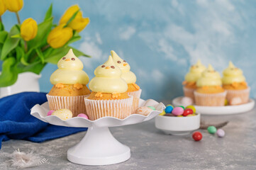 Cute Easter chick cupcakes. Vanilla cupcakes with buttercream on a white stand with fresh flowers on a white wooden background. Copy space.