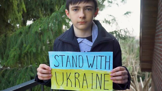 Poor boy kid homeless protesting armored conflict holding banner with inscription message text Stand with Ukraine on background of yellow blue flag. Crisis, peace, stop aggression in whole world