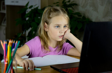 A little sweet girl undergoes online school education behind her laptop at home in the living room.