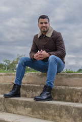 full length shot of a Colombian man smiling and looking at the camera while is sitting on a steps of a concrete stairs with a cloudy sky behind him