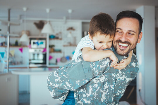 Happy reunion of one young soldier with family, five years old son and father having fun in the house during the beautiful sunny day. Soldier dad hugs his son after a time in military.