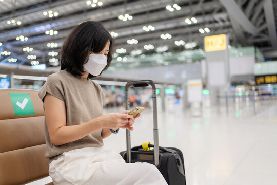 Woman  with luggage sitting on the chair and checking phone in the international airport terminal and wearing hygiene face mask to prevent Covid 19 outbreak. Transportation and new normal concept