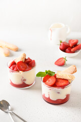 Layered dessert in glass jars with cookies, mascarpone cheese and whipped cream on white background. Italian tiramisu with strawberries.