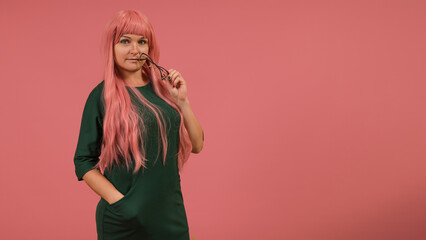 pretty young woman with long pink hair and a green dress with glasses in her hands on a pink background with copy space