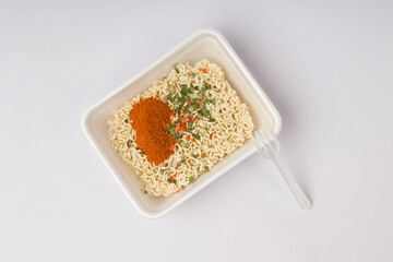 dry instant noodles with a plastic fork, vegetables and hot spices in a container on a white background. fast food concept