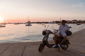A modern senior businessman in casual clothes sitting on a scooter by the sea and enjoys the sunrise. Selective focus 