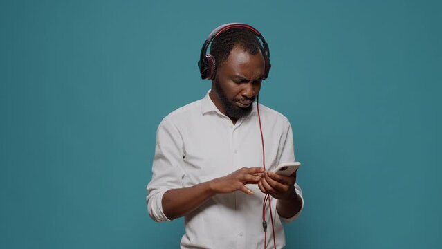 Young person listening to sound on headphones with mobile phone, paying attention to podcast. Confused man feeling uncertain about internet information on smartphone and audio heaset.