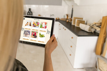 Woman shopping food online using a digital tablet at the kitchen, close-up view on a tablet screen. Concept of buying online using mobile devices.