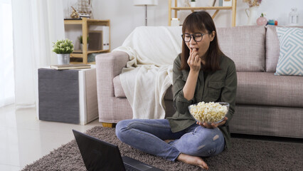 Obraz premium relaxed asian young lady streaming comedy series on notebook and enjoying popcorn in the cozy living room in daytime