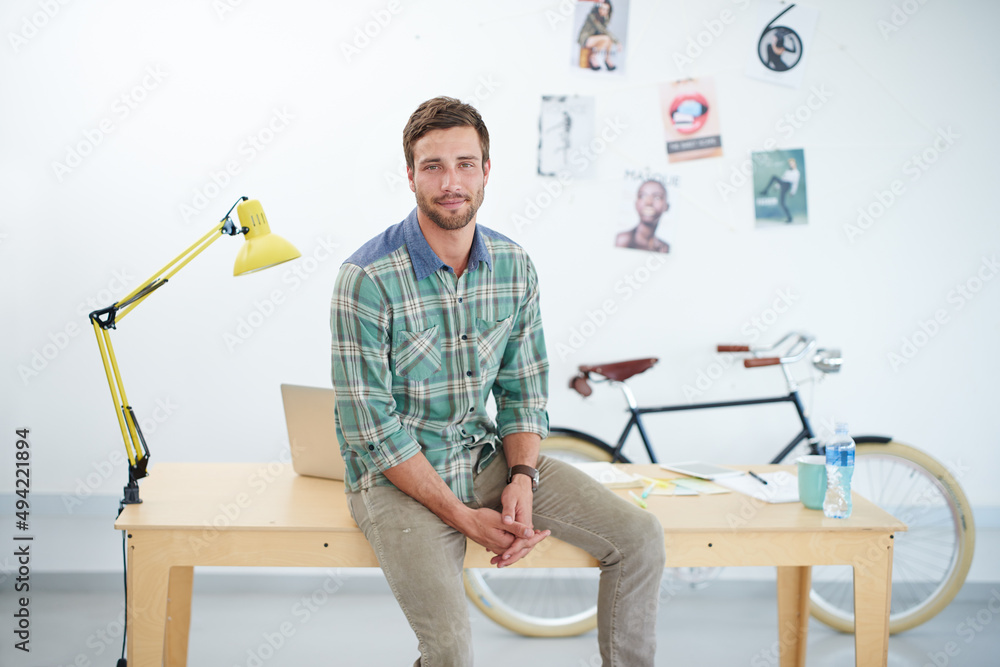 Poster portrait of a casually-dressed young man sitting on his desk in an office. the commercial designs di