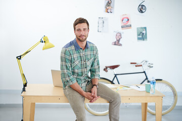 Portrait of a casually-dressed young man sitting on his desk in an office. The commercial designs displayed in this image represent a simulation of a real product and have been changed or altered