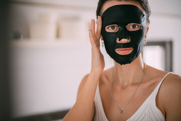 A beautiful brunette woman makes facial skin care procedures with a cleansing, moisturizing mask near the mirror in a bright room