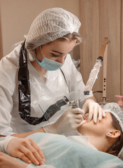 beauty procedure. process permanent lips tattoo. master in white disposable cap, medical gloves and mask makes permanent lip tattoo on a brunette model. lifestyle concept, free space