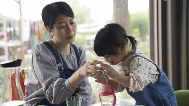 cheerful asian mother giving her cup and smiling while looking at her toddler daughter learning to drink water by herself in restaurant during breakfast time