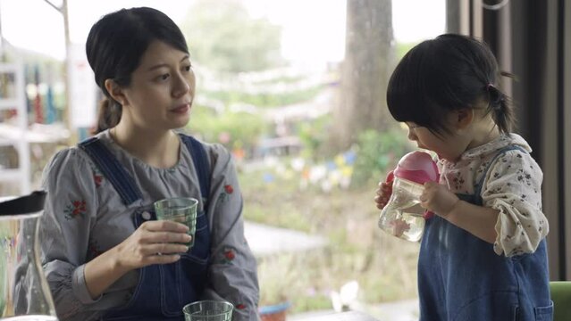 asian mother holding a cup is giving her toddler daughter sipper bottle to drink water with her in a restaurant with daylight at lunchtime.