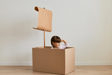 Portrait of charming little girl sitting in cardboard box images she is sailing a boat looking far....
