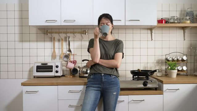 front view of a serious asian woman leaning on kitchen platform and looking into distance with a thoughtful face while drinking coffee in the morning at home.